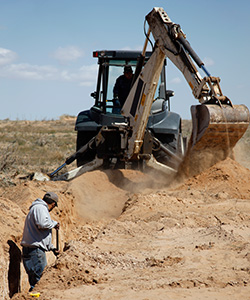 A worker digs in dusty a trench as a backhoe dumps dirt behind him.