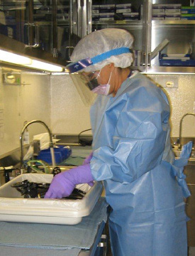 A worker wearing a gown, hair covering, full face covering, and gloves washes her hands in a sink.