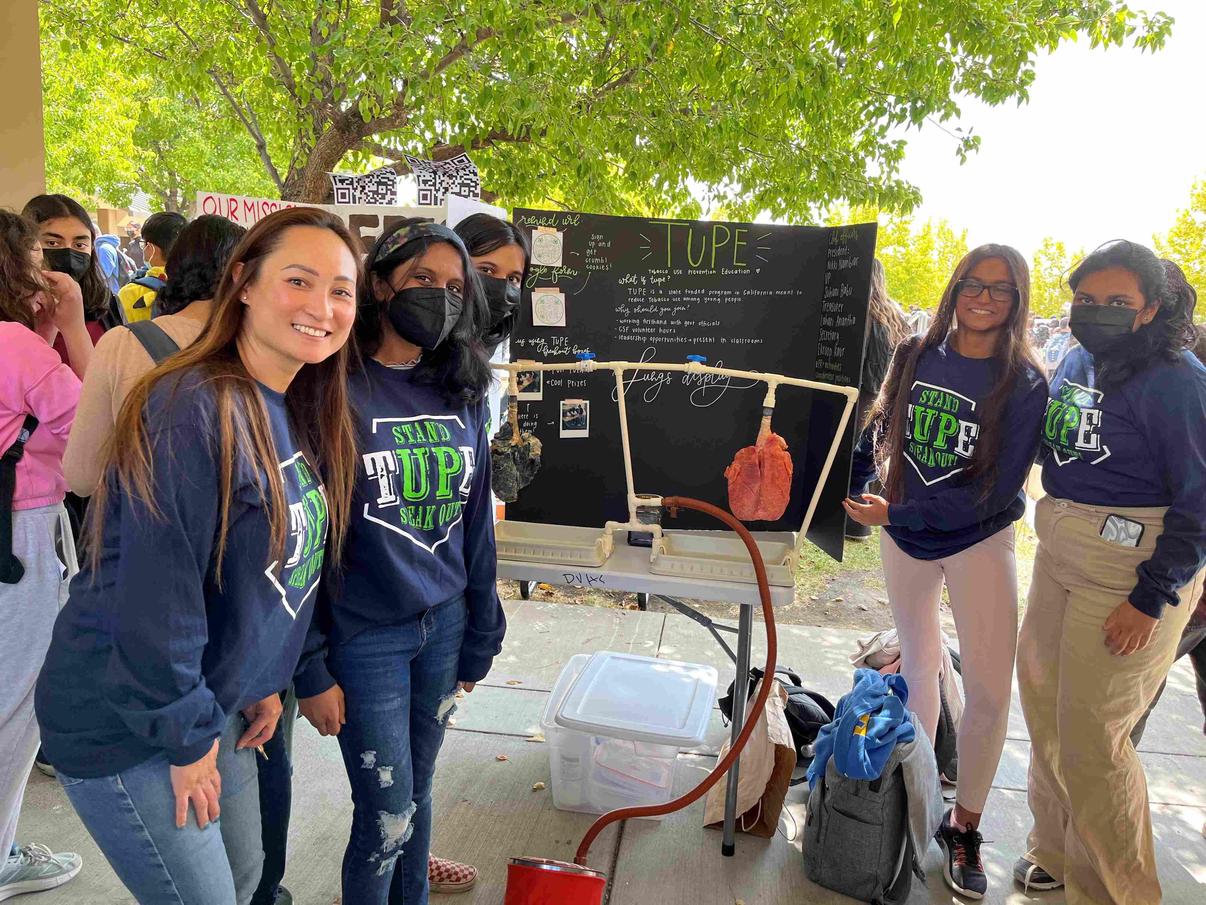 Five smiling high school students wear sweatshirts with the words “TUPE/Stand Up Speak Out.” Between them a table display shows 