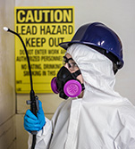 A worker with full protective gear, including goggles and a respiratory face mask, holding a blowtorch next to a lead hazard warning sign
