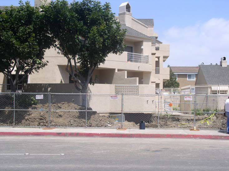 Exterior of a tan stucco apartment complex with angled terraces and walls, surrounded by a chain link fence to protect sidewalk pedestrians from an excavated pit of dirt around the side of the apartment.