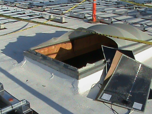 A skylight is raised slightly from a white rooftop and half the plastic covering is broken. There are jagged edges where the covering has broken and flat, black, flexible solar panels are placed on the roof near the opening.