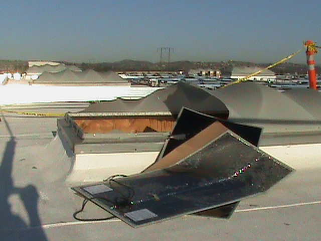 Two flexible black panels lie against a skylight with half the plastic covering missing. A yellow piece of tape surrounds the broken skylight.