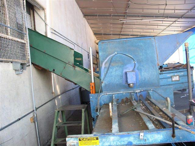 A short step-ladder stands under a chute leading from a building to a large, blue metal machine.