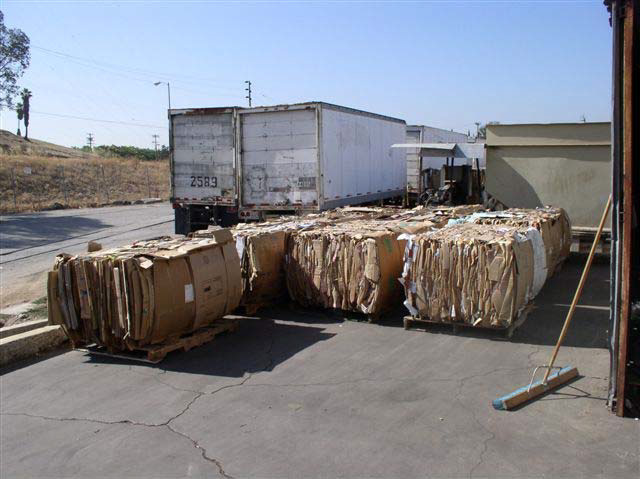 Stacks of compressed cardboard stand on asphalt next to two large truck trailers.