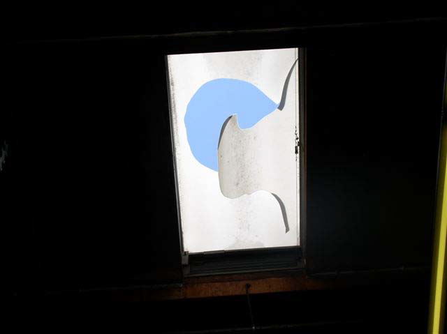 Blue sky can be seen from a broken skylight that has a piece of material hanging from a jagged opening in the center.