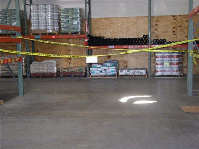 Yellow tape is tied across two large shelving units in a warehouse to block access to an area. There are bags, pipes, boxes, and tubs stacked in pallets against the wall behind the roped-off area.