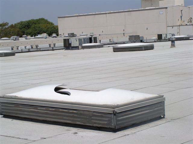 A broken skylight is on a white rooftop. In the distance are more skylights and a rooftop air conditioning unit.