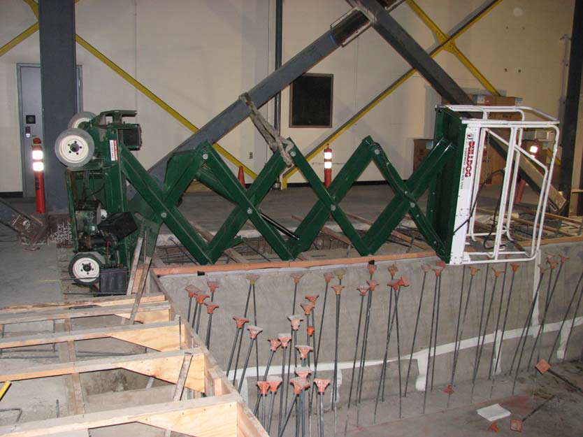 A fully extended scissor lift lies on its side directly above a concrete pit with metal spikes rising from the bottom of the pit.