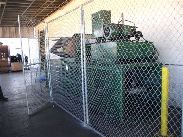 The side of a large, green metal baling machine behind a chain link fence.