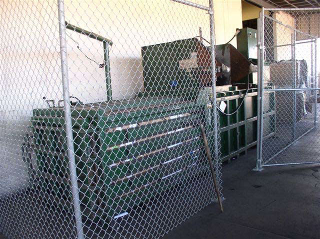 The left side of a dark green, metal baling machine behind a chain link fence.