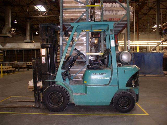 A blue-green forklift is parked in front of a tall shelving unit inside a warehouse. The front wheels are larger in diameter than the rear wheels.
