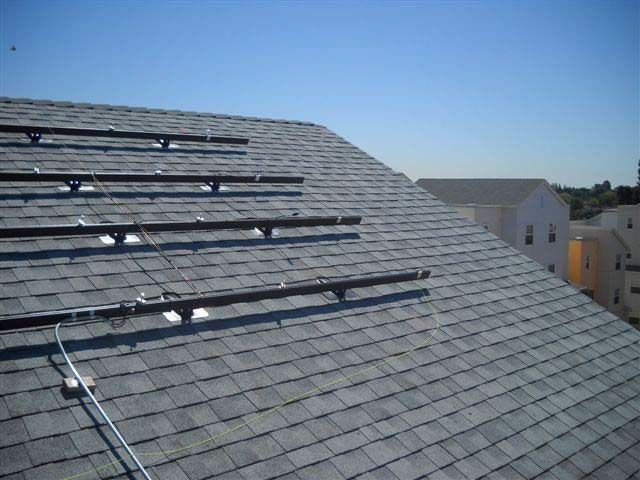 Four horizontal mounting brackets for solar panels are attached to the steeply slanted roof of a building, and there are other multi-story residential buildings in the distance.