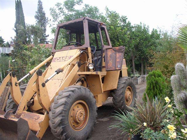 A large yellow tractor with a shovel in the front. It is high off the ground with large tires and a small opening with a rusted door to the driver's cab.