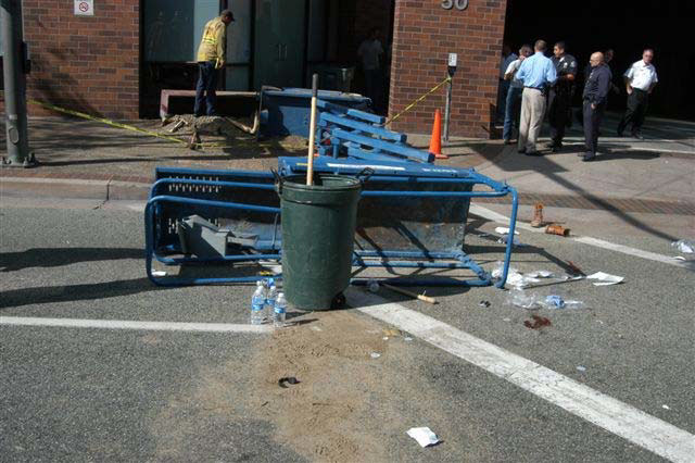 An upright garbage pail stands in front of the platform of the toppled lift. Blood and dirt are on the street next to the platform.