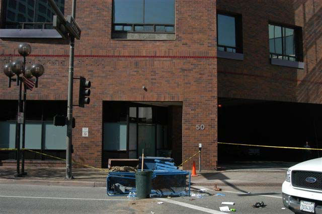 The fallen lift lies on a street in front of a multi-floor dark-red brick building. The lift is in front of an entrance and a window on the second floor of the building.