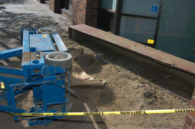 The bottom of the overturned lift with its wheels exposed is surrounded by dirt and rocks from the fallen planter beside it.