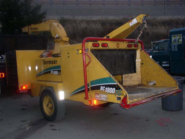 A yellow Vermeer BC 1800 XL wood chipper on wheels with a flat loading area in the back sits parked in a lot.