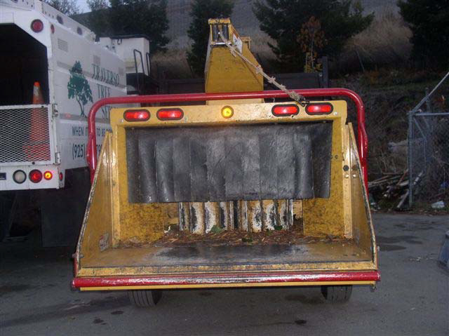 A view of the back of the wood chipper with a flat metal loading area that leads through a black ribboned half curtain to the chopping blades. There are four red reflector lights on the top of the back of the wood chipper.