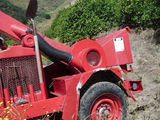 The side of the wood chipper with a tool holder, which has a shovel placed upright in it. The metal around the wheel arch is dented and the paint is missing.