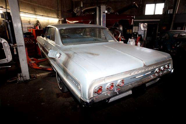 Rear end of a white 1960s Chevrolet Impala seen from the left rear side. The fuel filler door is open and there are black burn marks around open fuel cap, the rear quarter panel, and on the trunk lid near the fuel filler door.