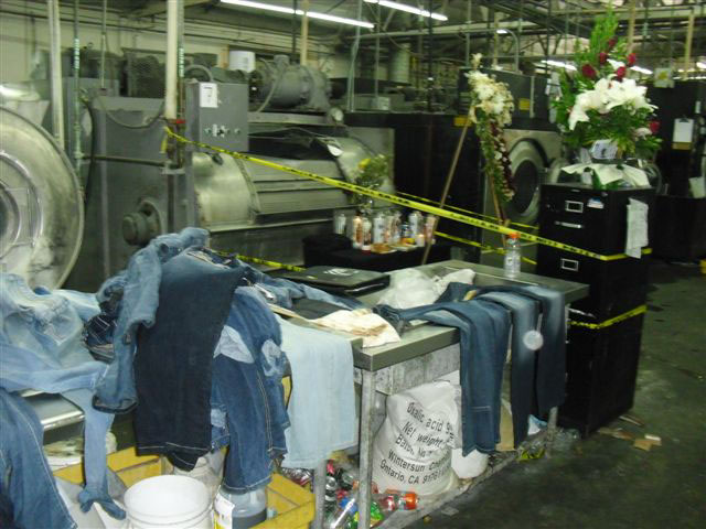 Denim clothing is piled on a work table near a large metal washing machine that is surrounded by yellow tape. Flowers are set on a filing cabinet nearby and a large floral wreath is in front of the machine where the worker died.