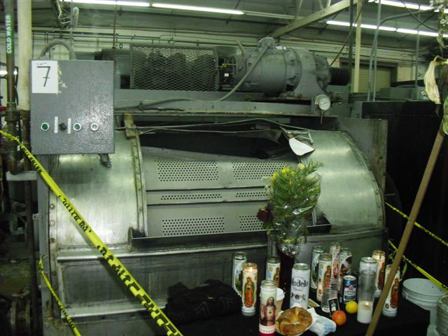 Candles and flowers sit on a table in front of the damaged metal industrial washing machine where the worker died. The machine has a damaged, bent semi-cylindrical sliding vented door opening where the worker became trapped.