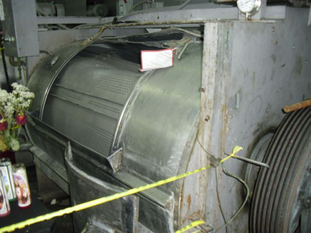 Close-up of the metal industrial washing machine with a large dent on top where colleagues tried to pull the trapped worker out of the machine. The machine is encased in metal with sharp corners.