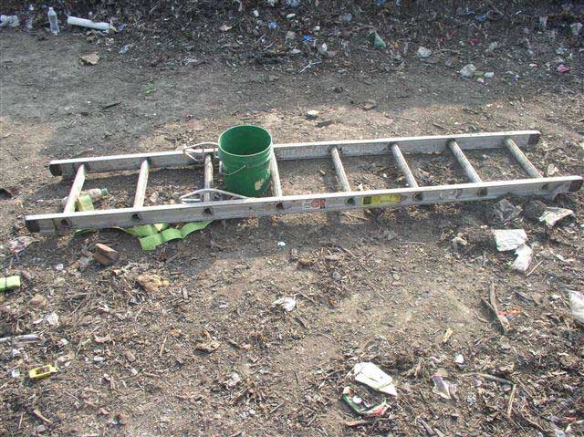 A green bucket stands between the third and fourth rungs of an 8-rung ladder that is lying on dirt surrounded by scattered trash.