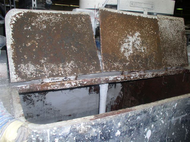 A paint-splattered tank sits in an industrial warehouse with three corroded metal panel lids in the open position.