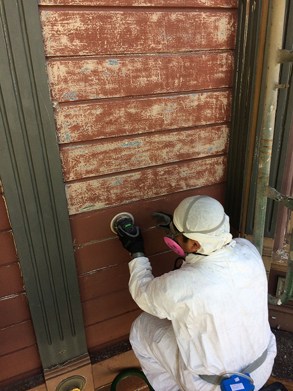 A worker wearing a white protective suit kneels while sanding paint off the exterior wall of an older building.