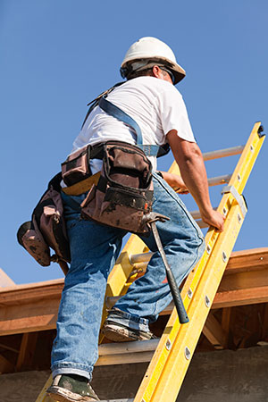 A construction worker wearing a hard hat and tool belt climbs a ladder onto a roof.