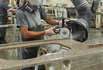 A worker wearing protective gear uses a saw with a water spray to cut a countertop while an industrial vacuum draws dust away.