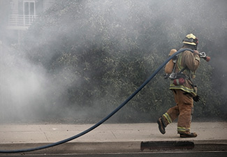 A firefighter carries a hose in the parking lot of a residential building amid heavy smoke.