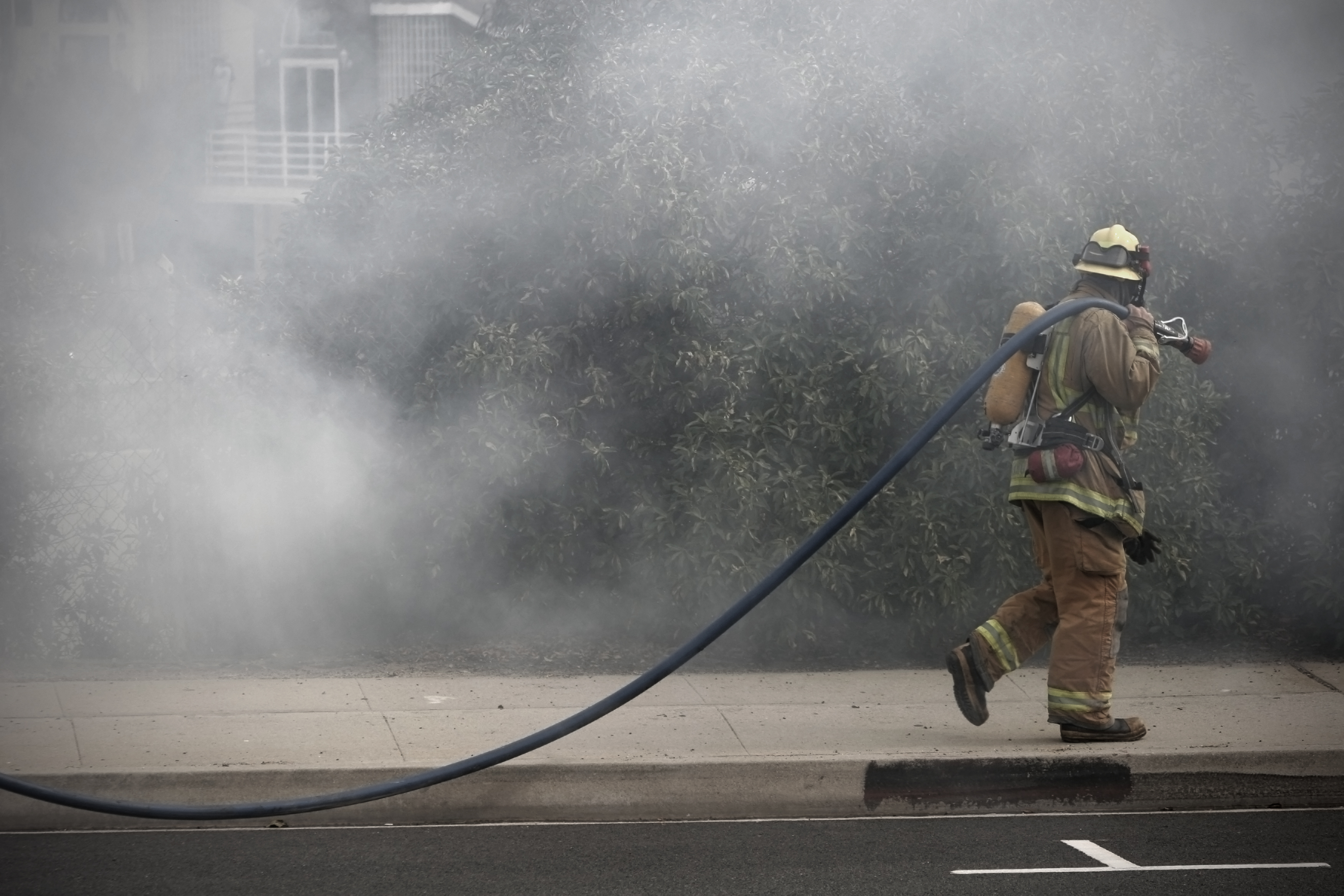 A firefighter walks with a hose near a sidewalk next to a smoky building.