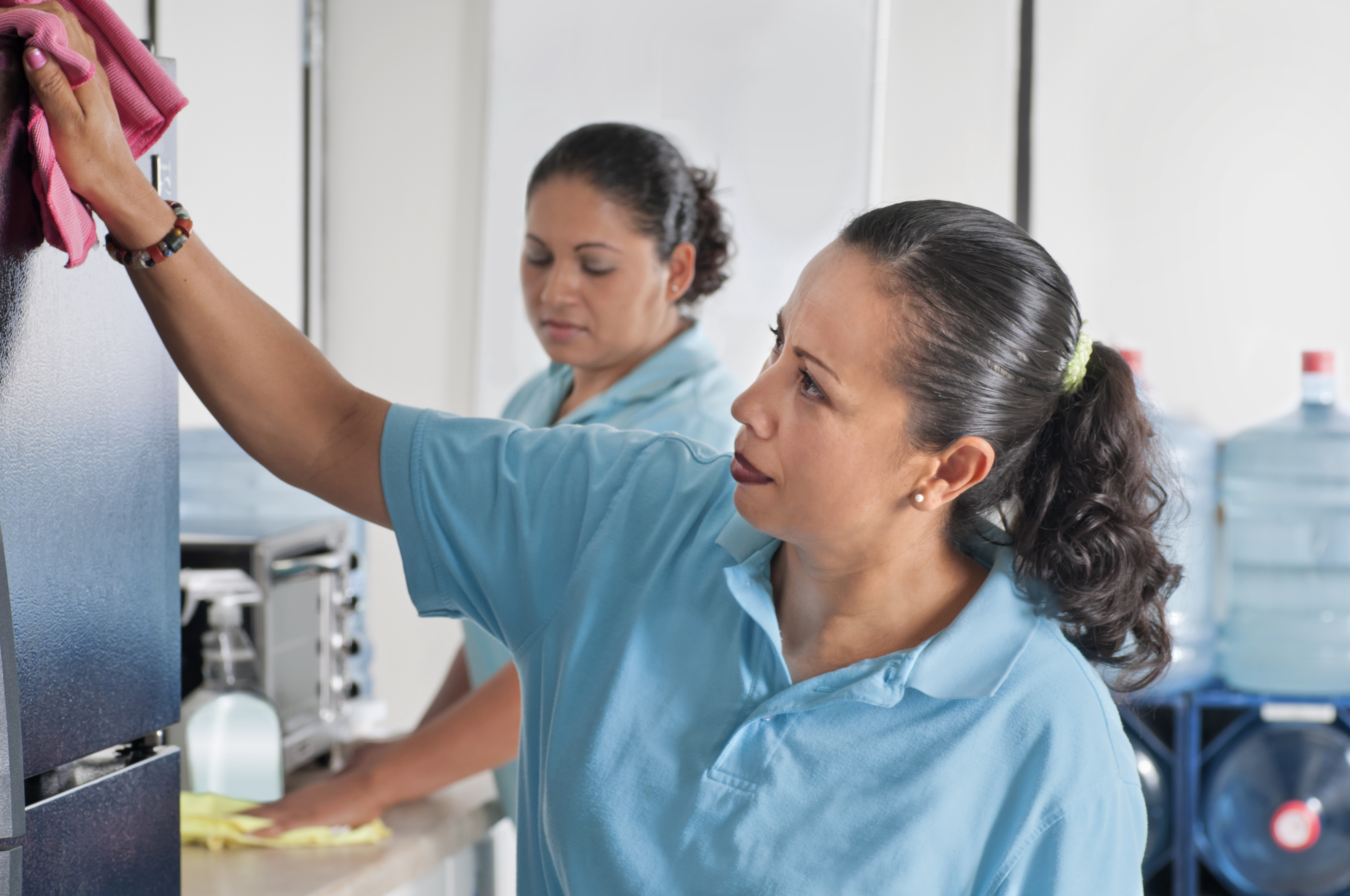 Two Latina women use microfiber cloths to clean the appliances in an office kitchen.