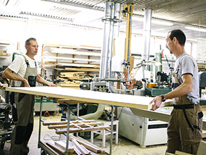 Two workers wearing protective clothing lift a heavy board in an industrial workshop.