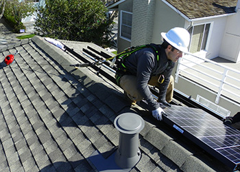 A man in a hard hat squats on a rooftop and holds a solar panel close to the roof.