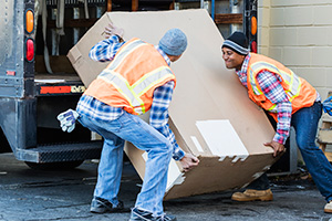 Two men struggle while lifting a heavy box from behind a large cargo delivery truck.