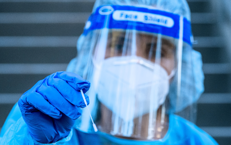 Healthcare worker wearing full PPE including a fitted respirator and face shield. She has a nose swab in her hand