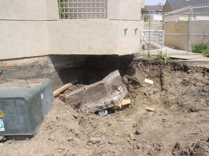 A large slab of concrete lies underneath an overhanging terrace in the bottom of a dirt pit that extends beneath the street level of an apartment building.